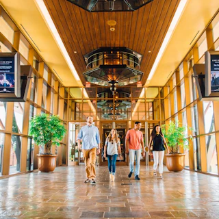 A modern, spacious lobby with large glass windows and wooden accents. Four people walk together, flanked by potted plants and digital displays.