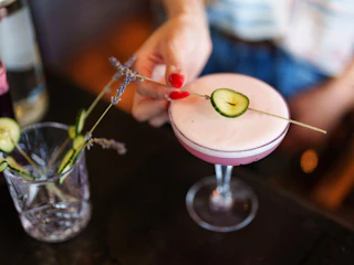 A hand garnishes a pink cocktail with a cucumber slice and lavender sprig, alongside a glass with similar garnishes.