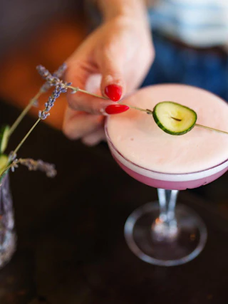 A hand garnishes a pink cocktail with a cucumber slice and lavender sprig, alongside a glass with similar garnishes.