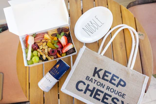 A wooden table displays a gourmet snack box filled with fruits, nuts, and a drink, alongside a plate labeled "NIBBLE" and a tote bag.