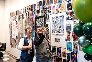Two men take a selfie in front of a vibrant wall covered with movie posters at the Baton Rouge Underground Film Festival.