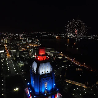 Aerial view of a city skyline at night, featuring a brightly lit tower in red, white, and blue, with fireworks in the background.