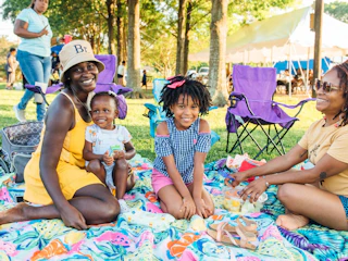 A family enjoys a picnic on a colorful blanket in a park, surrounded by trees and folding chairs. Laughter and snacks create a joyful atmosphere.