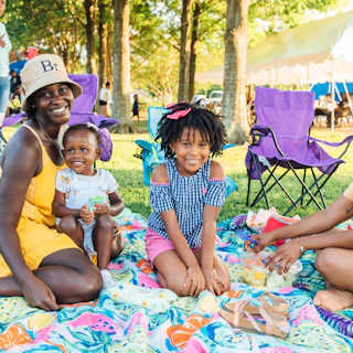 A family enjoys a picnic on a colorful blanket in a park, surrounded by trees and folding chairs. Laughter and snacks create a joyful atmosphere.
