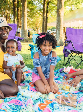 A family enjoys a picnic on a colorful blanket in a park, surrounded by trees and folding chairs. Laughter and snacks create a joyful atmosphere.