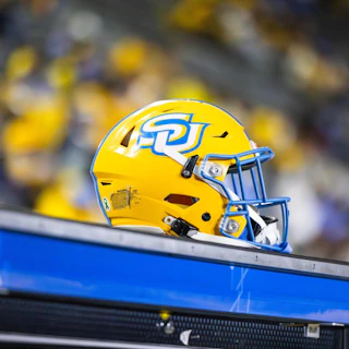 A bright yellow football helmet with a blue logo sits on a bench, surrounded by a blurred crowd in the background. The scene captures the excitement of a game day atmosphere.