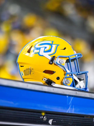 A bright yellow football helmet with a blue logo sits on a bench, surrounded by a blurred crowd in the background. The scene captures the excitement of a game day atmosphere.
