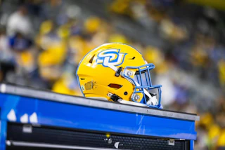 A bright yellow football helmet with a blue logo sits on a bench, surrounded by a blurred crowd in the background. The scene captures the excitement of a game day atmosphere.