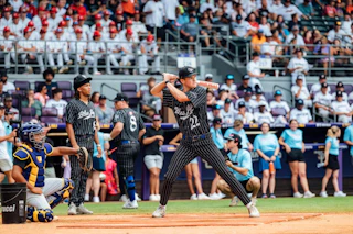 A baseball player stands ready to bat on a field, surrounded by teammates and a cheering crowd in the stands. The scene captures the excitement of a live game.